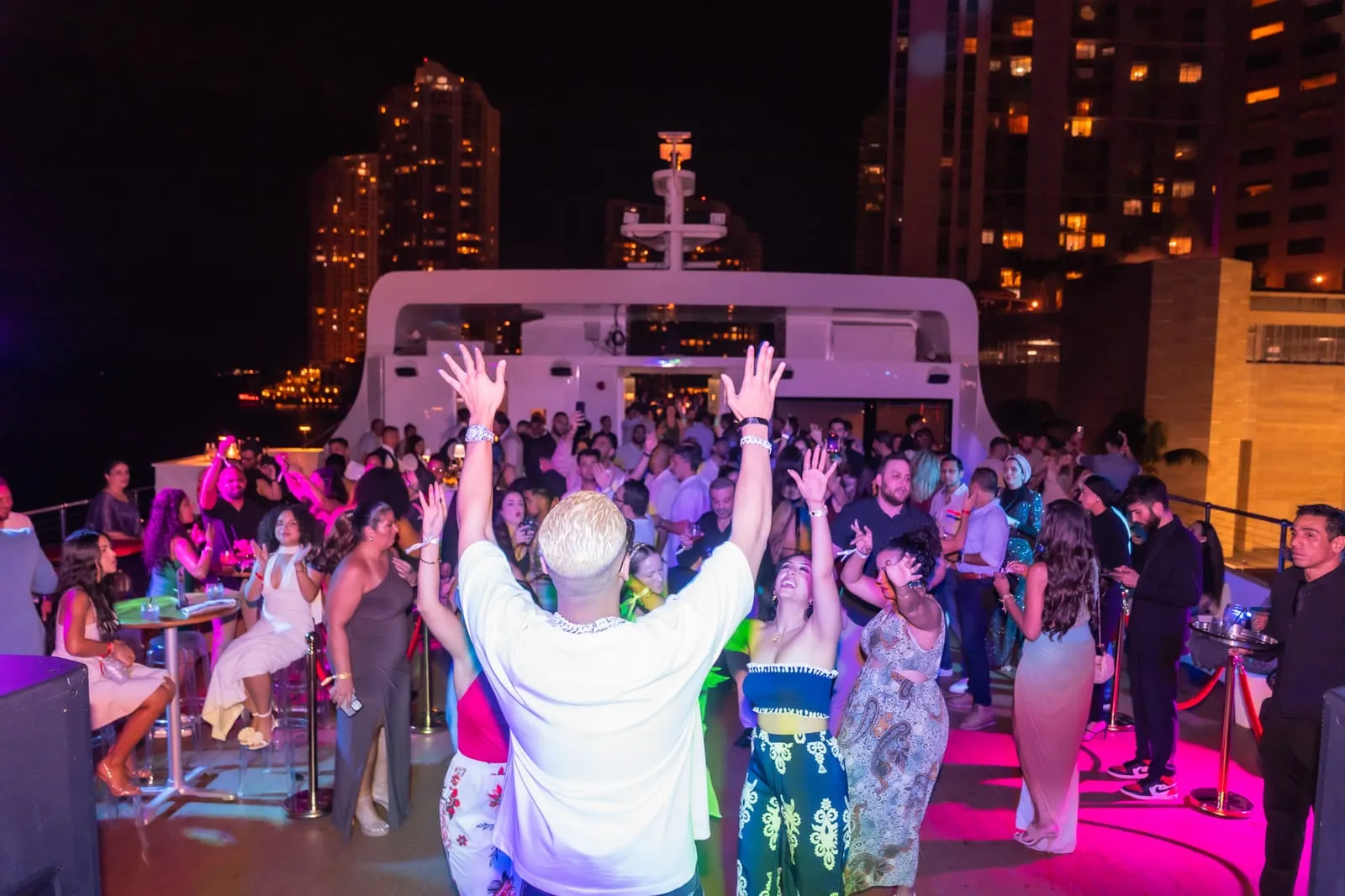 Guests dance with hands raised on the deck at the Habibu Hub yacht gala, city towers lit up behind